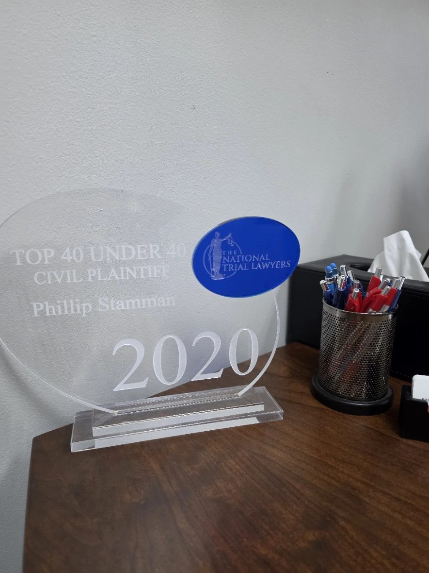 A clear acrylic award reading Top 40 Under 40 Civil Plaintiff, Phillip Stamman, 2020, from The National Trial Lawyers, on an office desk beside a cup of pens