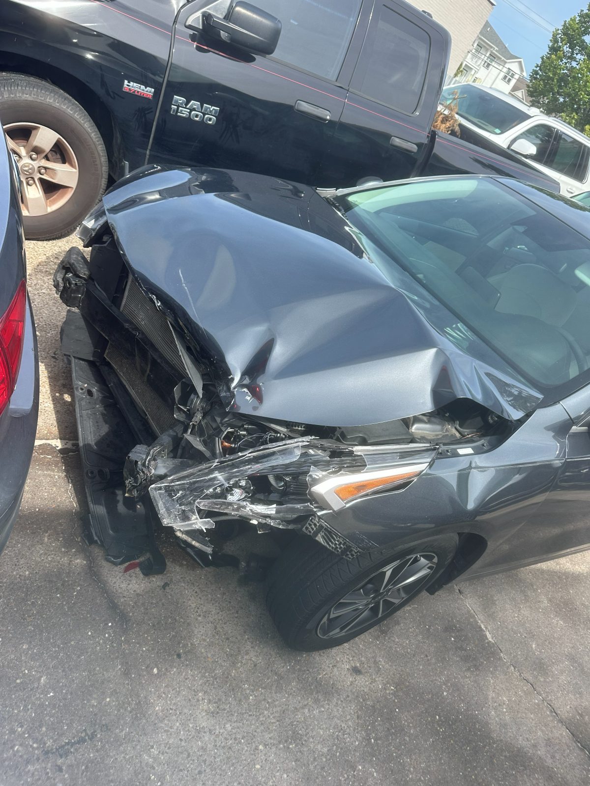 A gray sedan with a crushed hood, broken headlight, and torn-off front bumper parked in a lot after a crash