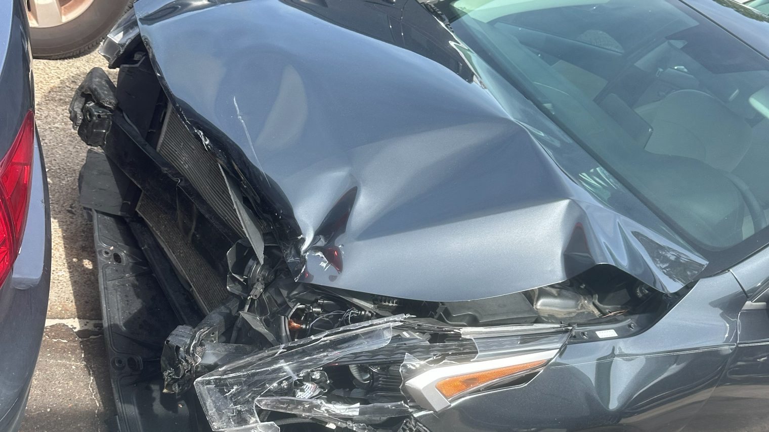 Front-end damage to a gray sedan after a collision, with the hood folded, the radiator exposed, and the headlight assembly torn away
