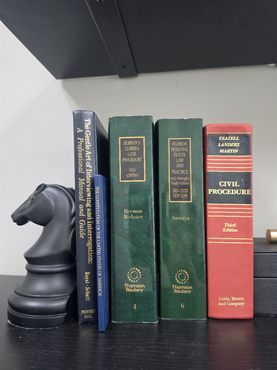 Law books on a black office shelf, including Florida Personal Injury Law and Practice and The Gentle Art of Interviewing and Interrogation, held up by a black chess knight bookend